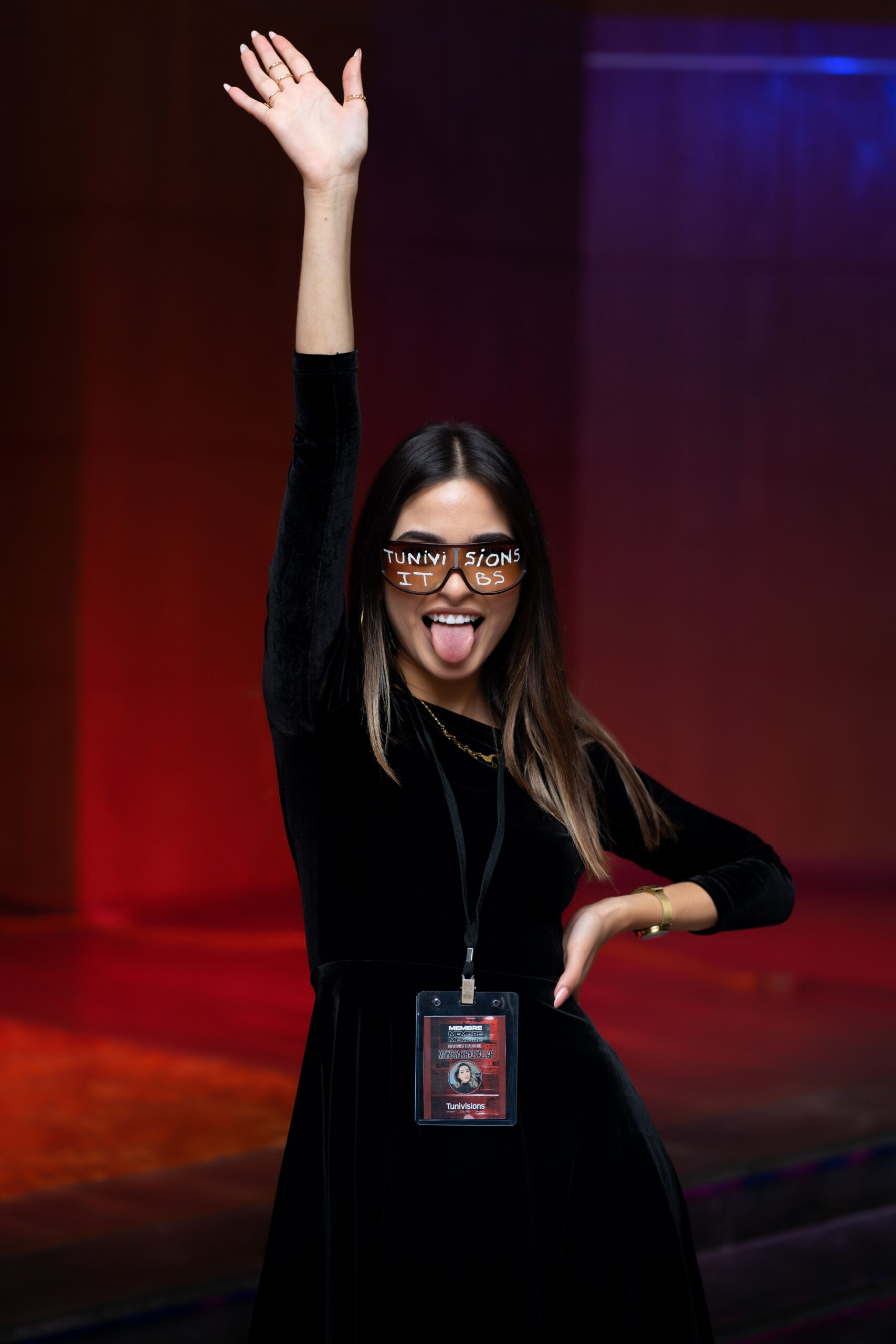 Young woman in black dress poses playfully with sunglasses and ID badge, enjoying a night event.